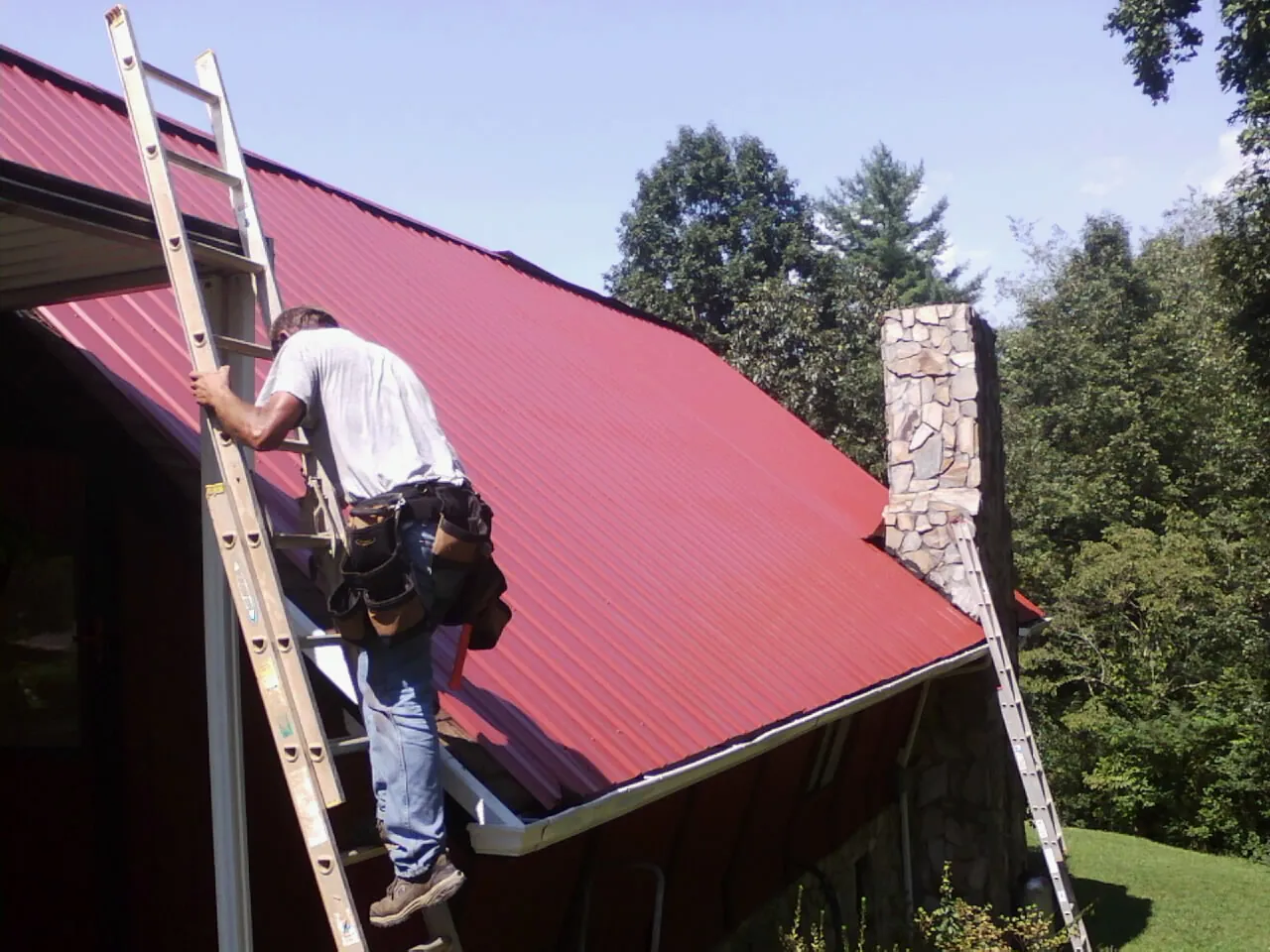 Roofer on a ladder beside a red metal roof and stone chimney — Soffit & Fascia Repair by Roofing Brothers in Moore